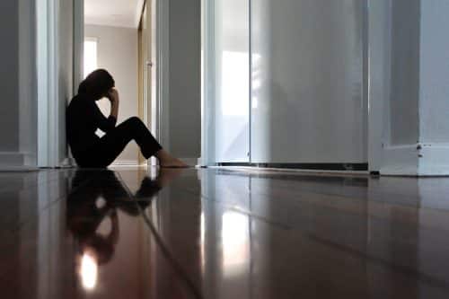 A distressed woman sits on the floor of her home with her head in her hands.