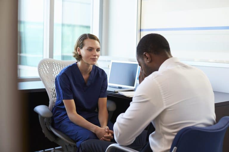 A doctor sits at her desk beside her patient whose head is in his hands.