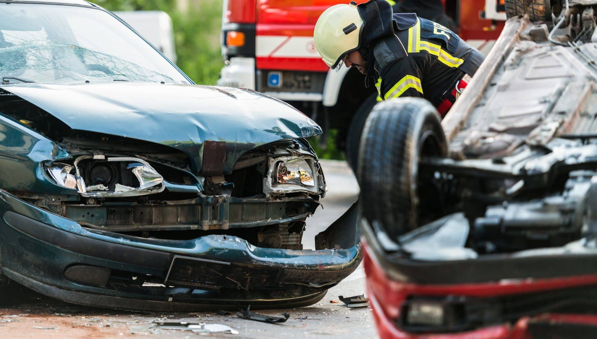 A first responder examines the front of a damaged vehicle after a car accident. An overturned vehicle lies behind him.