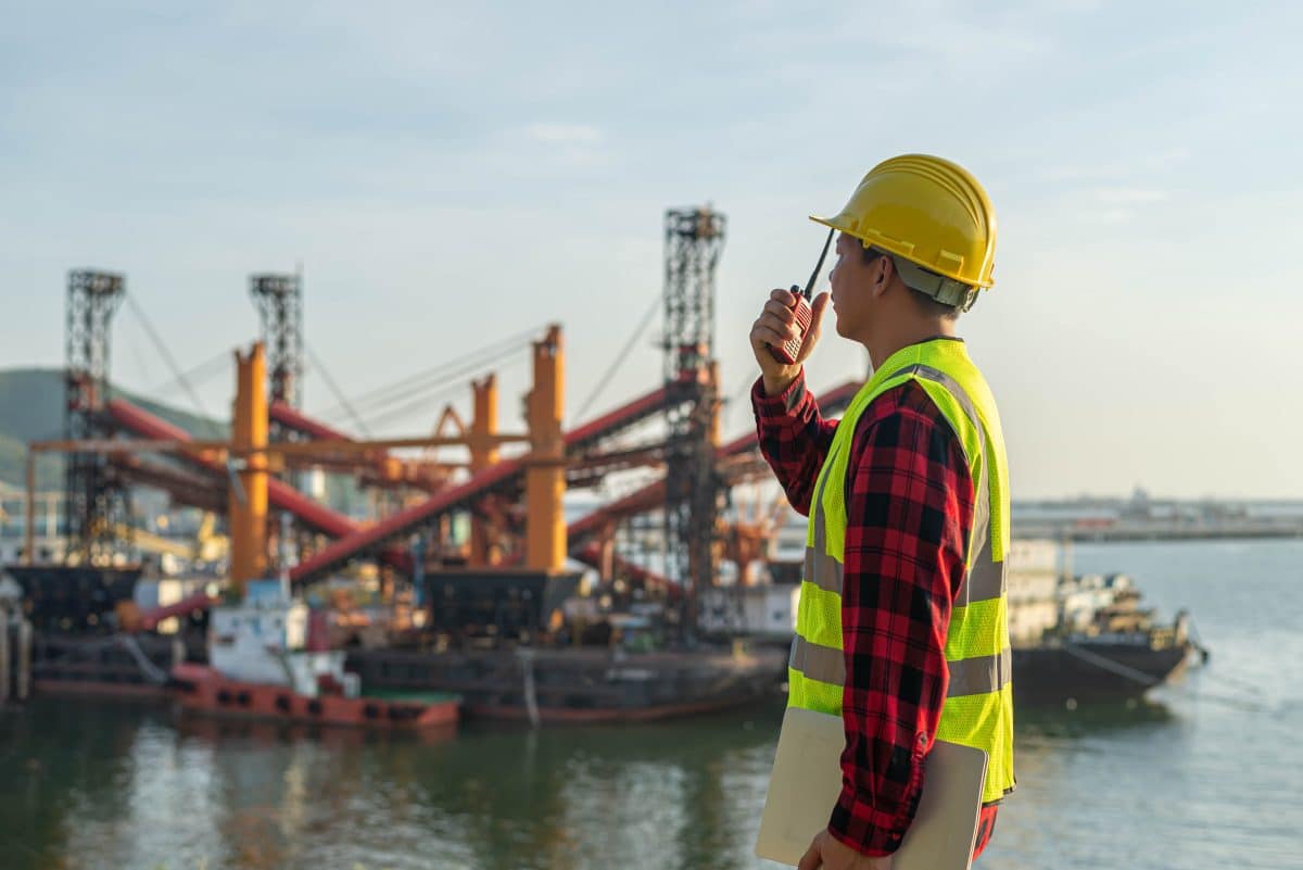 Man wearing a construction vest and hard hat talks into a walkie-talkie while looking out at a boatyard.