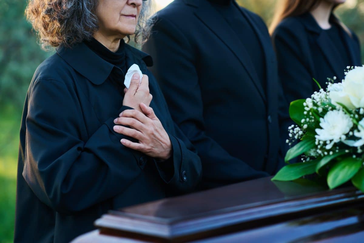 A mourning woman in black stands beside other mourners in front of a casket with flowers on it.